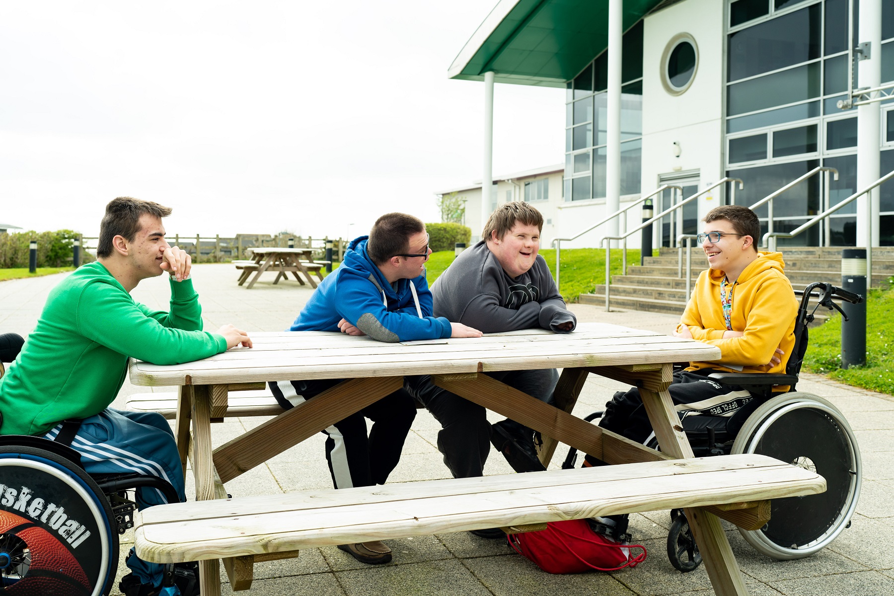 A group of students sit outside at a picnic bench chatting and smiling. Two of the students sit in wheelchairs.