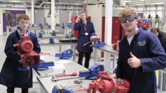 Three of the new engineering apprentices in blue overalls give thumbs up to camera while working on red machinery in desk clamps in the college's engineering workshop.