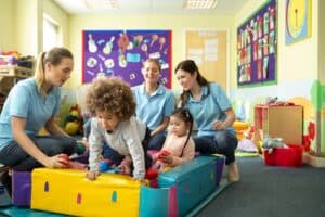 A horizontal image of a group of children in a ball pool playing and laughing, behind them sit three nursery teachers in blue polo shirts supervising and smiling. The nursery is a colourful scene of toys and mess.