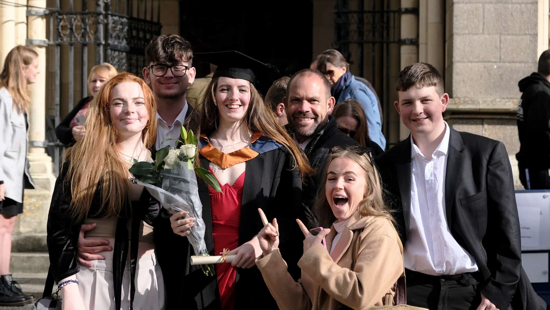 Family And Friends 2 For Web A graduate stands on the steps outside Truro cathedral with a bunh of white roses and lilys with her family around her smiling down camera. One of the graduates guests is pointing at her while cheering.