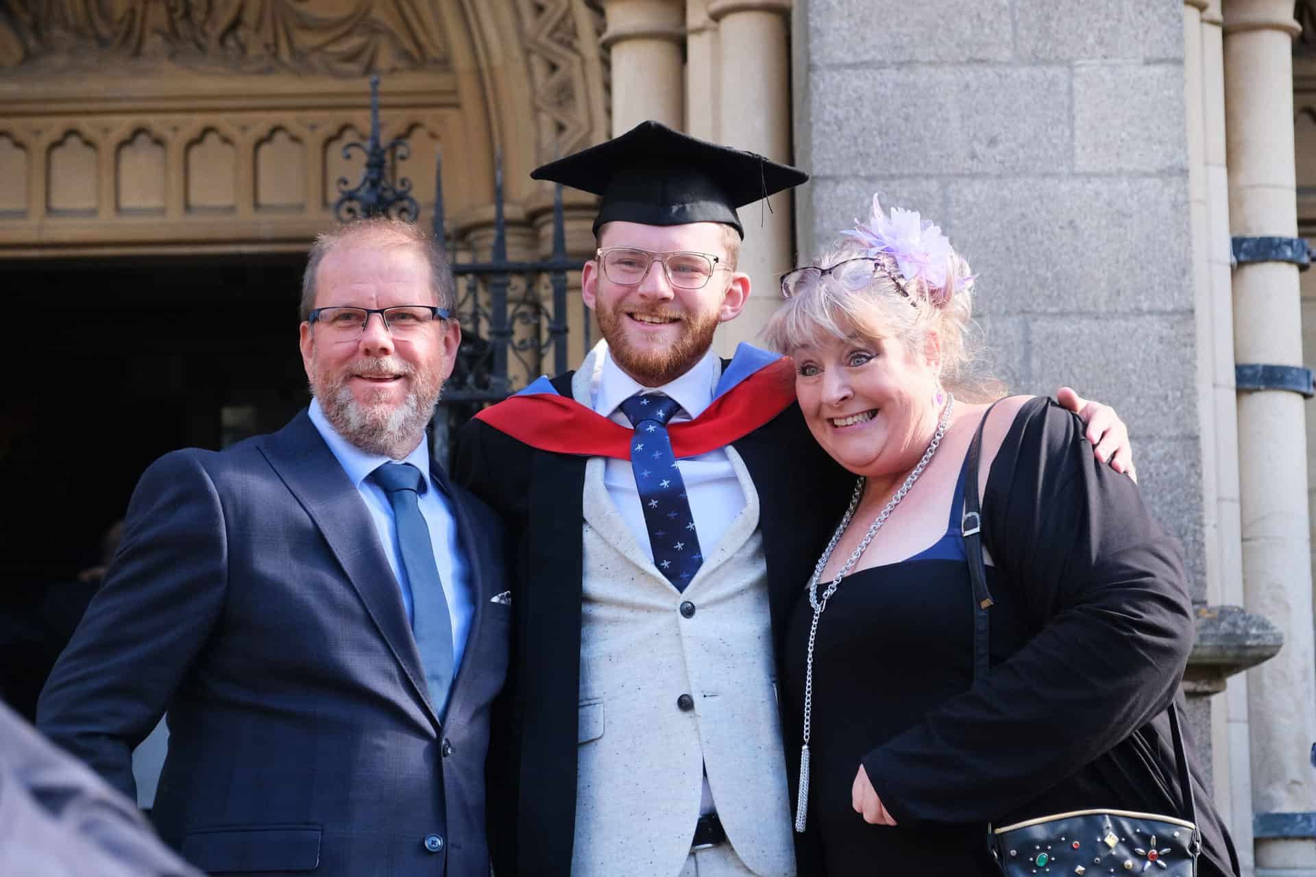 Graduate 13 The University Centre Truro & Penwith Graduation 2023. A graduate stands smiling outside the cathedral with his arms around two guests.