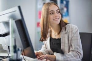 Smiling young woman sitting at the desk in her home office setup working on a project on her computer, looking over towards the living room with a bright smile towards her family. Freelance Working - Working from Home Business Concept.
