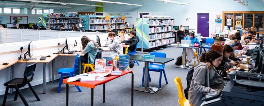 Students sit in the Learning Centre at Truro College. There are computers and bookshelves.