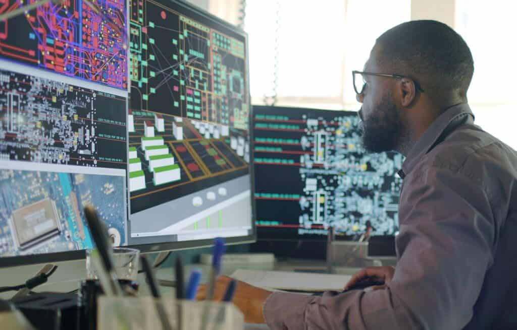 Image of a male designing electronic circuit boards. He’s sitting at a desk with a large computer screen displaying a variety of apps and illustrations.