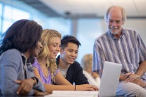 A group of students gather around a laptop in a classroom, smiling happily, while their teacher looks over at their work.