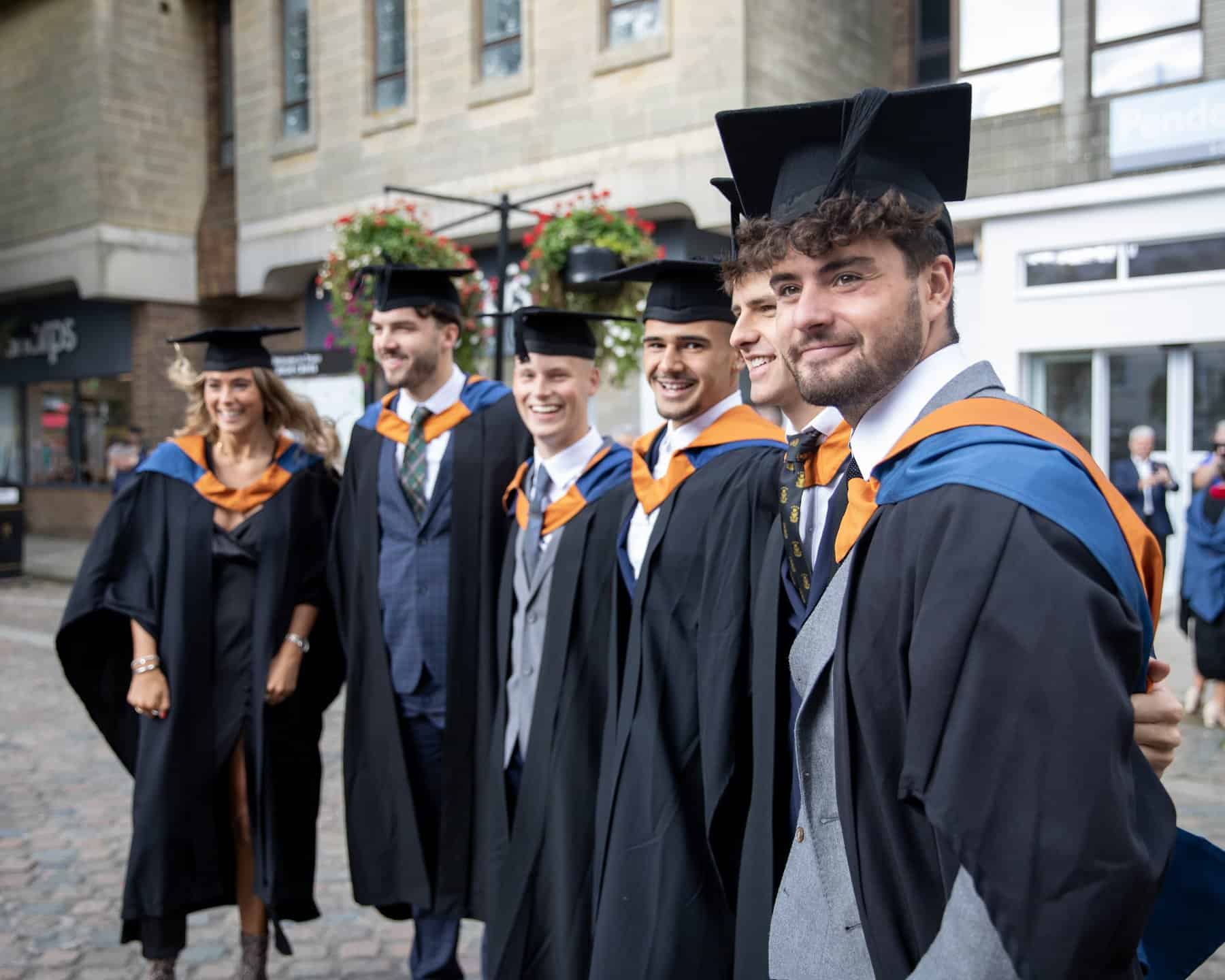 A group of Graduates in caps and gowns stand in a line with their around around each other outside Truro Cathedral.