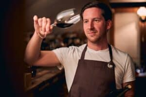 Confident young adult in a brown apron standing in a winery cellar and looking at a glass of wine while tilting it.