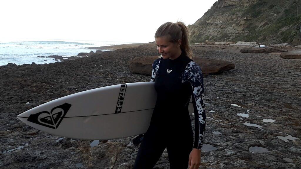 Surfer Lauren walks across the beach with her surf board under her arm.