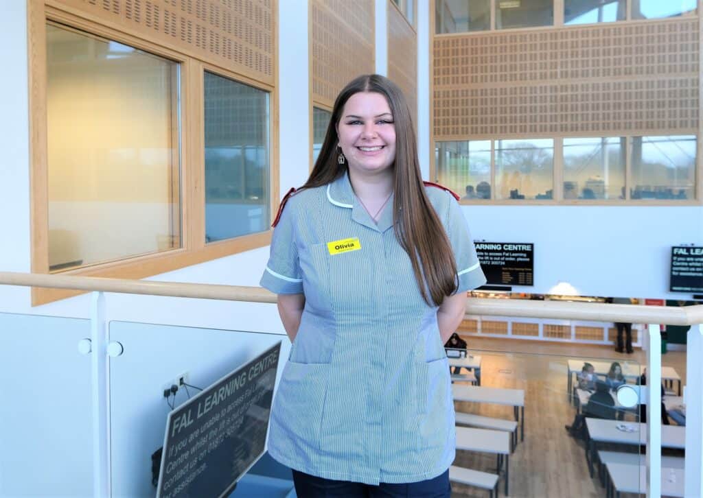 a Student nurse with long brown hair who is wearing a black and white striped uniform with red shoulder patches smiles in the University Centre Fal building.