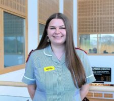 a Student nurse with long brown hair who is wearing a black and white striped uniform with red shoulder patches smiles in the University Centre Fal building.