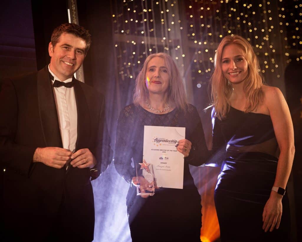 Academic Mentor of the Year winner Tamzin Irvin holds her awards on stage while stood between Holly Day and Scott Mann MP, spotlights shine and star lights twinkle in the background.