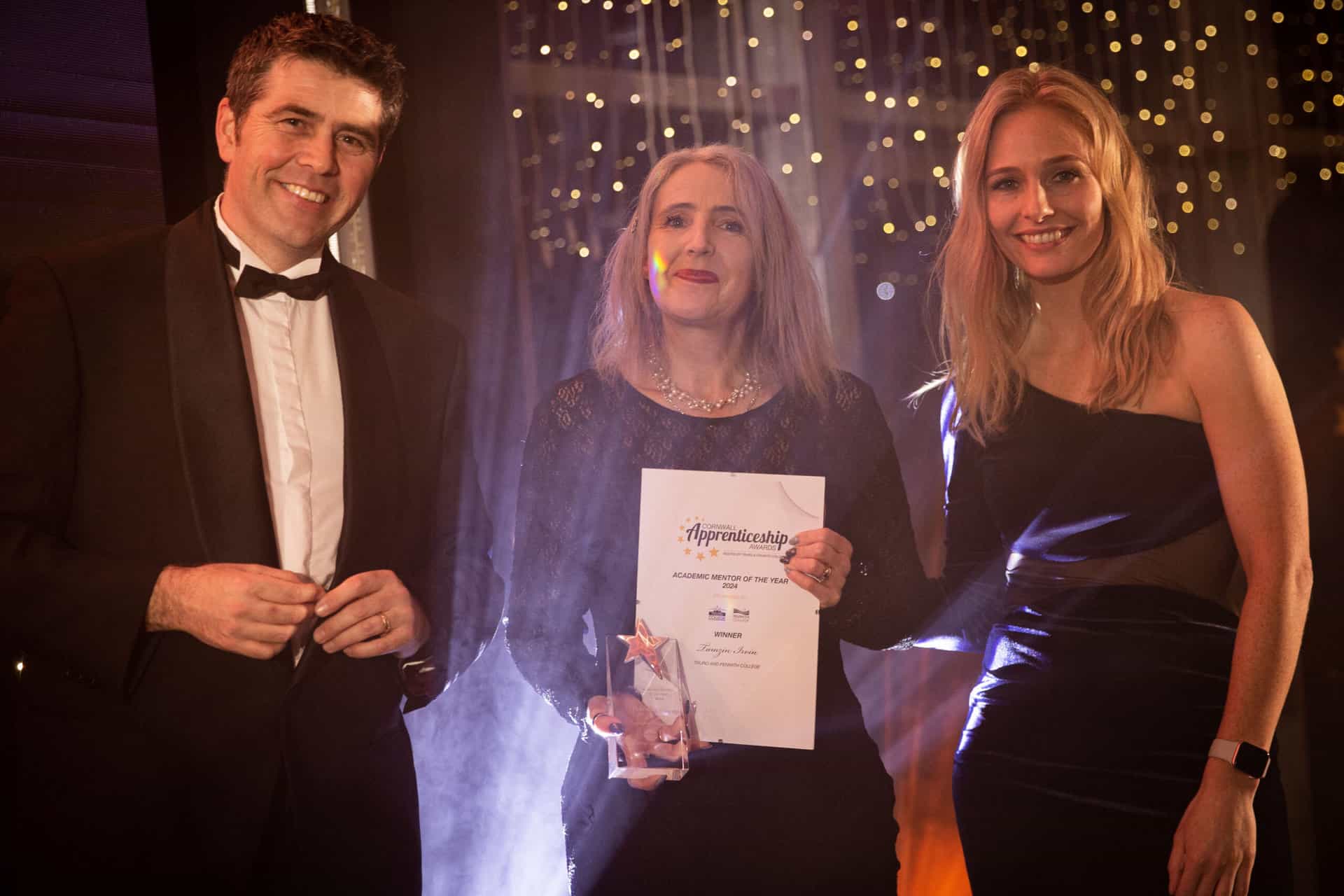 Academic Mentor of the Year winner Tamzin Irvin holds her awards on stage while stood between Holly Day and Scott Mann MP, spotlights shine and star lights twinkle in the background.