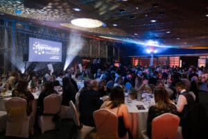 A panorama shot of the Awards in Eden Project's Gallery room. The lights are down with spotlights shining. Over 200 guests are sat at 22 round tables with stage at the back of shot with Awards logo on the large 4 meter screen.