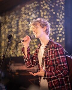 Music student Jake sings live on stage during the reception, holding a microphone to his mouth with twinkly star lights in the background.