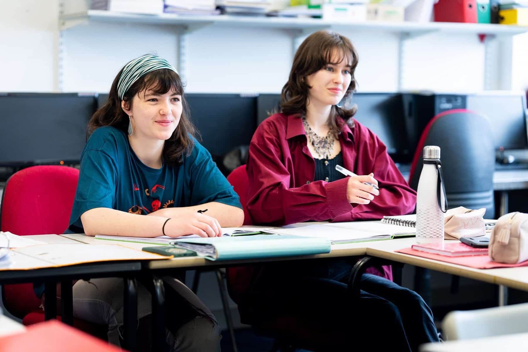 Two students sit at a classroom table in Penwith College. They have pens in hand and notebooks open in front of them as they face towards a teacher who is not in view.