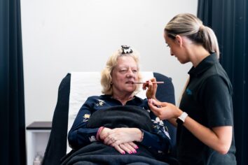 A beauty student is applying lipstick to a client's lips as they are sat comfortably in a reclined position.