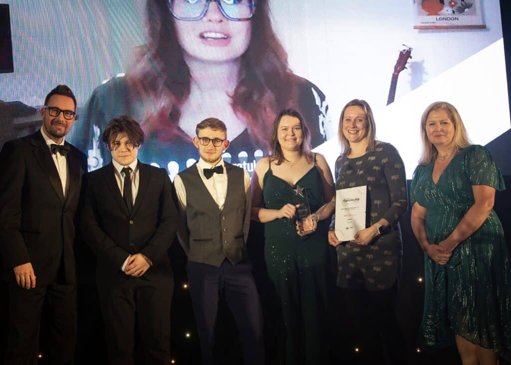 Four members of staff from Hiyield pose for a group photo on stage with their glass trophy and certificate. Neil Caddy from Pirate FM is far left with sponsor presenting the Award Cathie Kessell far right.