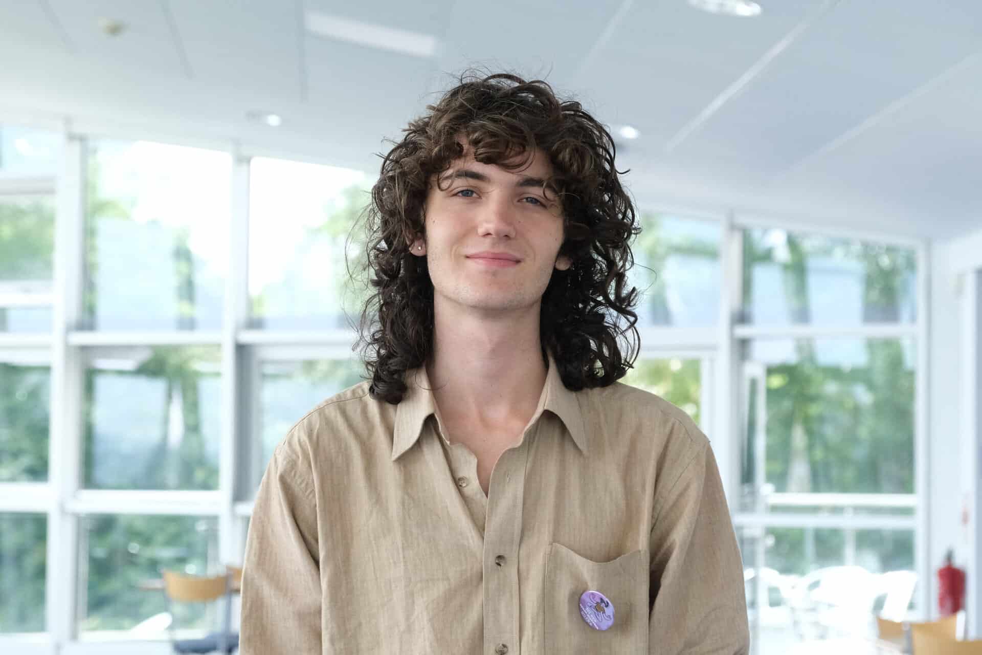 Kit A Level English Literature, History and Politics at Truro and Penwith College. Kit is wearing a light brown shirt and is stood in a sun-lit room smiling at the camera.