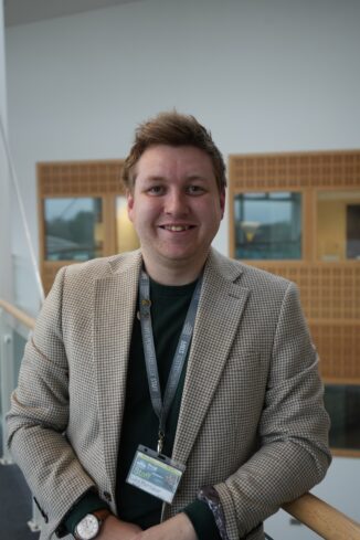 An image of Luke, an apprentice alumni of Truro and Penwith College. Luke is wearing a blazer and stood on the balcony section of the Fal building, smiling at the camera.