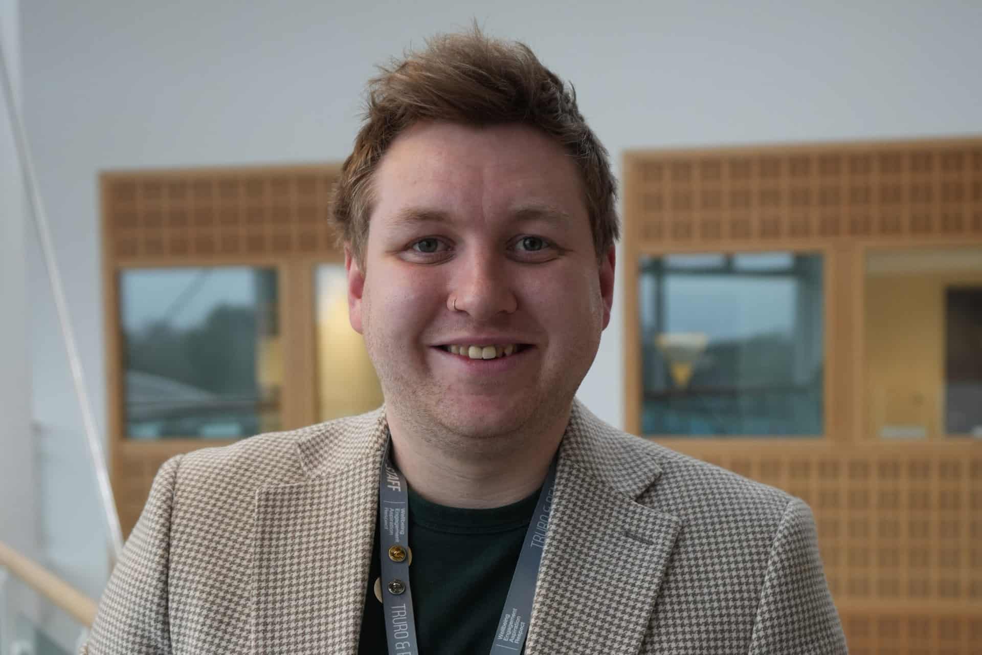 An image of Luke, an apprentice alumni of Truro and Penwith College. Luke is wearing a blazer and stood on the balcony section of the Fal building, smiling at the camera.
