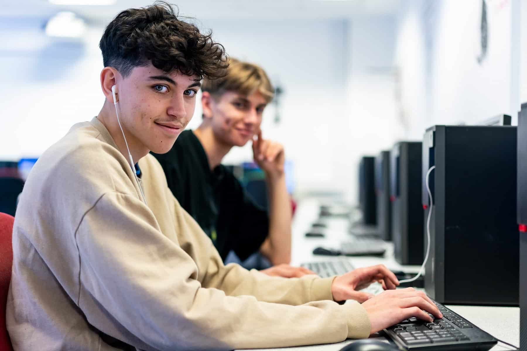 A student with earphones in turns to look at the camera. He is sat in front of a computer.