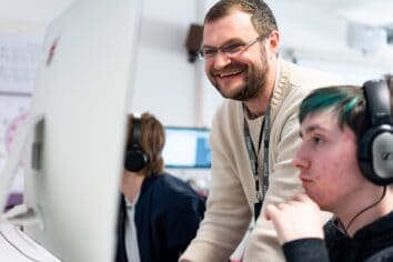 A tutor leans towards a student's computer screen in a helpful way. He is smiling.