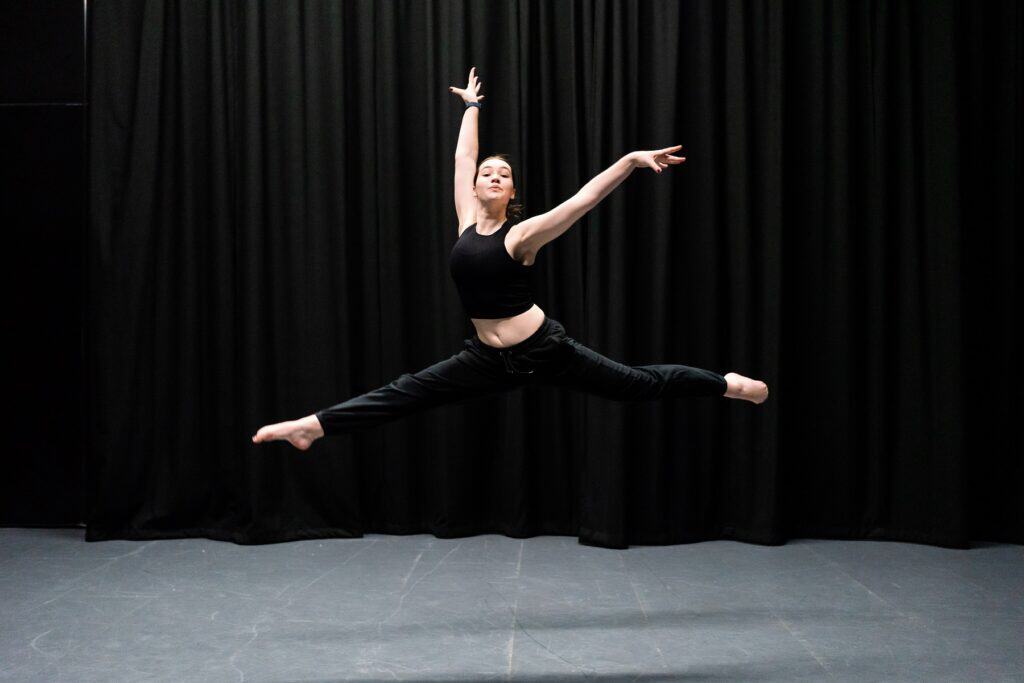 A performing arts dance student leaps into the air on a stage with a dark backdrop.