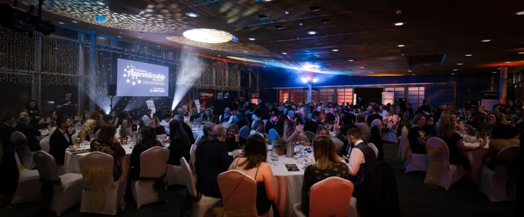 A panorama shot of the Awards in Eden Project's Gallery room. The lights are down with spotlights shining. Over 200 guests are sat at 22 round tables with stage at the back of shot with Awards logo on the large 4 meter screen