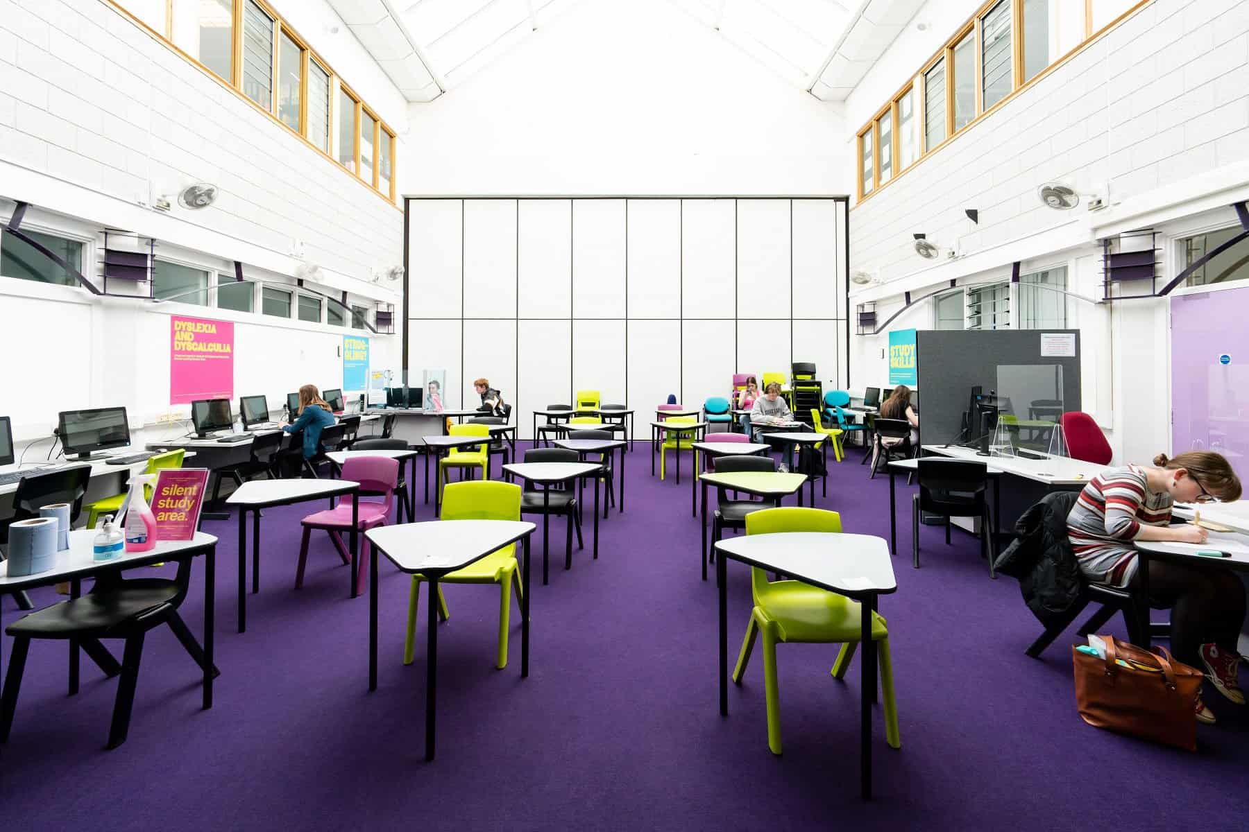 An image of the quiet study centre at Truro College. There is purple carpet and a mix of desks and chairs layed out.