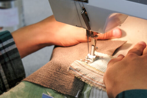 The camera zooms in on hands working a sewing machine. They are sewing pieces of brown and green materials together.