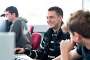 A student sits at a computer desk with a mouse in his hand. He smiles towards the screen while another student in foreground is turned towards him.