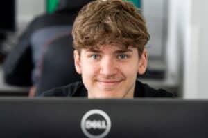A student looks over the top of their computer screen towards the camera and smiles.
