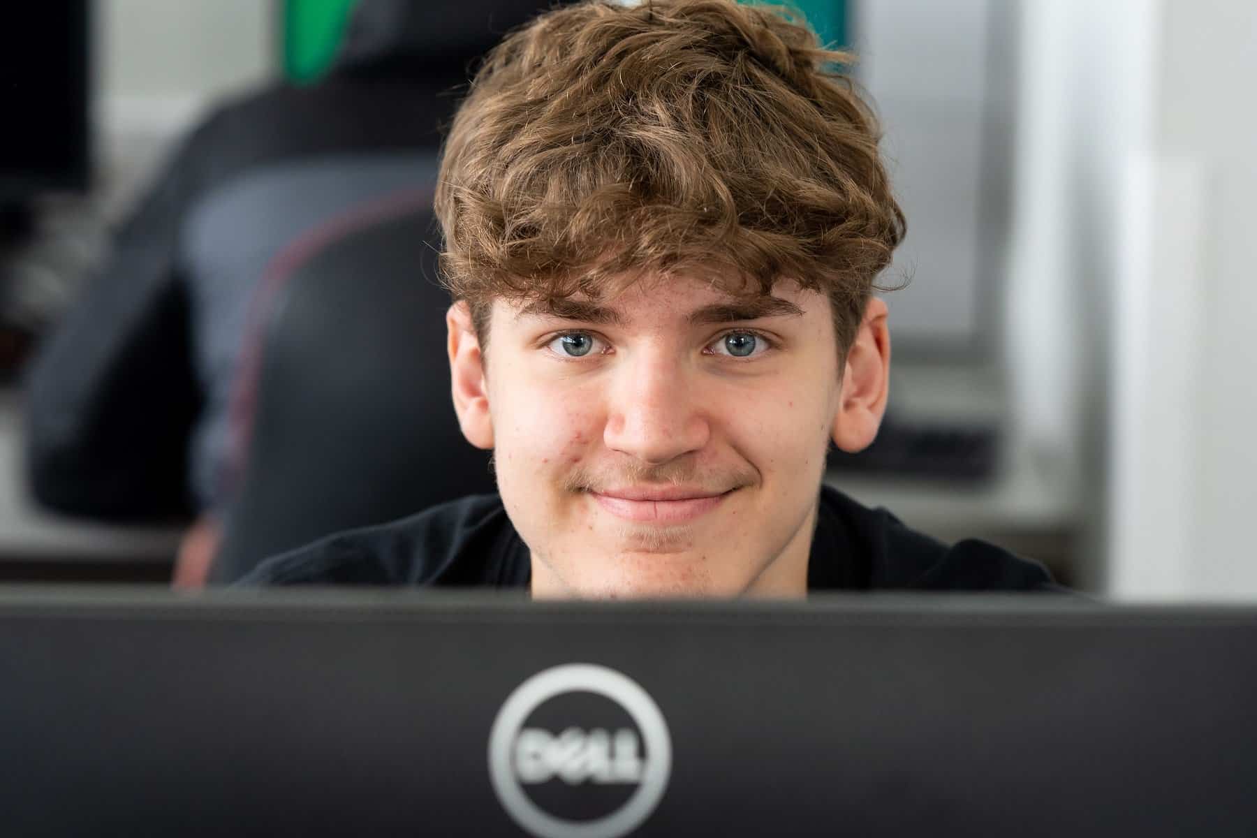 A student looks over the top of their computer screen towards the camera and smiles.