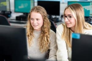 Two students sit side by side looking at the same computer screen. They both have long blonde hair and one is wearing glasses.