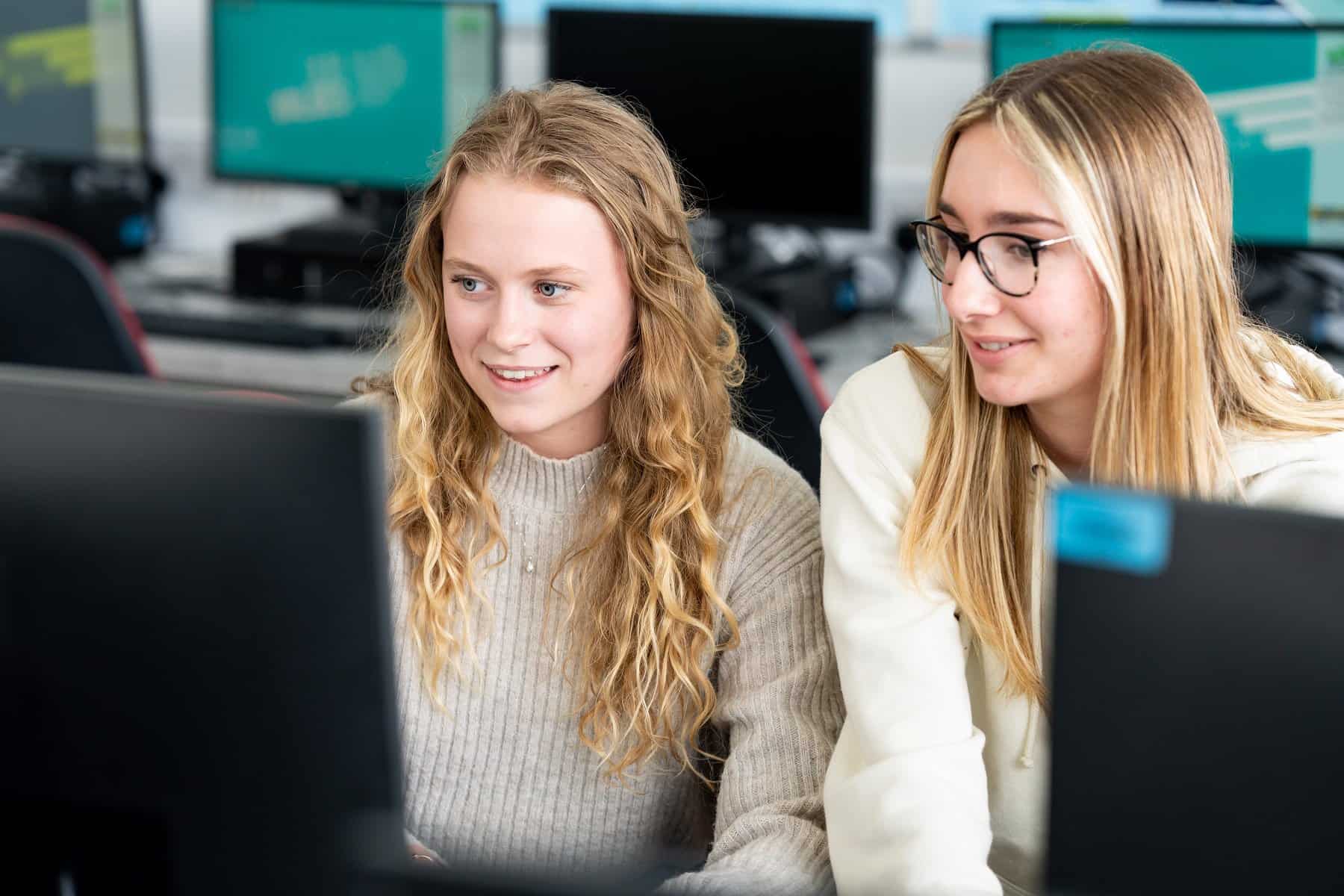 Two students sit side by side looking at the same computer screen. They both have long blonde hair and one is wearing glasses.