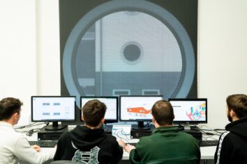 The camera looks over the shoulders of four students who each have a computer in front of them. They are looking at building designs. There is also a large circular window in front of them.