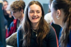 A student with long brown hair is smiling at another student who is unknown. They are wearing a dark jumper and gold jewellery.