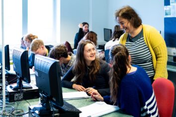 A computer room of students. Two students look up at their tutor who is smiling at them.