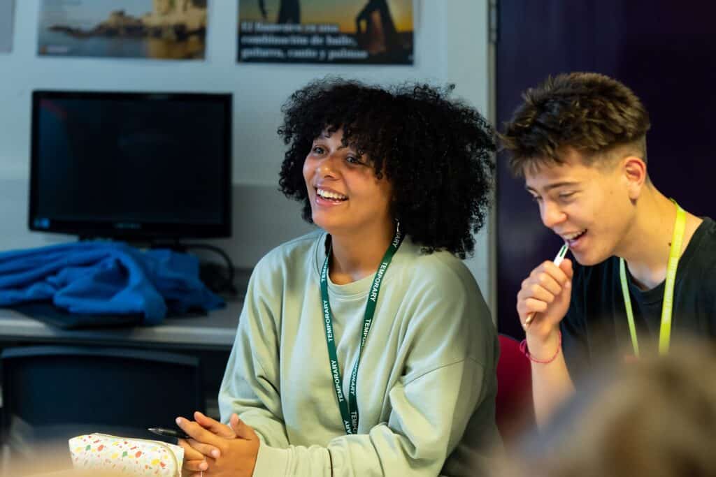 Two students sit in a classroom. A student with dark curly hair and a green jumper smiles towards someone we can't see. A pencil case in front of them.