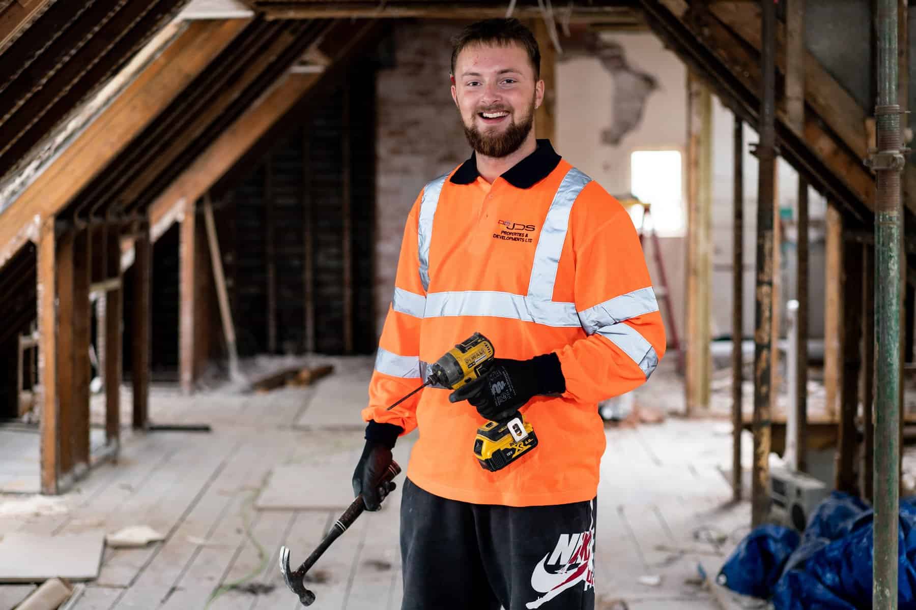 A young man in high visibility clothes holds a hammer and electrical tool while smiling at the camera. He stands in a room currently being renovated.