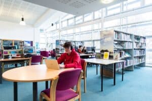 A student sits in the library at Penwith campus surrounded by bookcases, desks and computers.