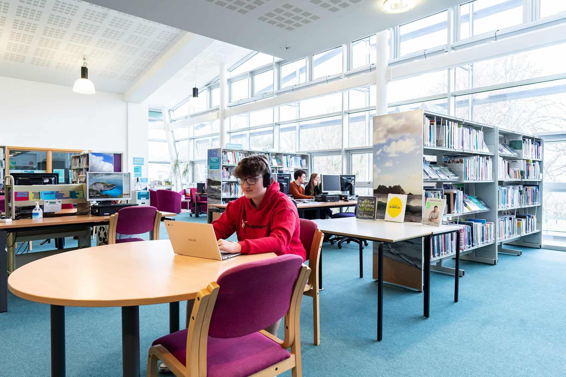A student sits in the library at Penwith campus surrounded by bookcases, desks and computers.