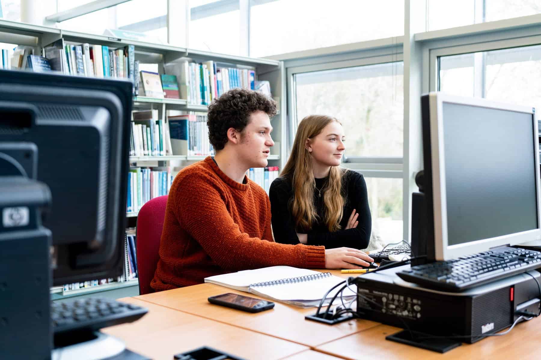 Two students sit in the library at Penwith College and look at a computer together. One student has their hand on the mouse. There are bookcases behind them.