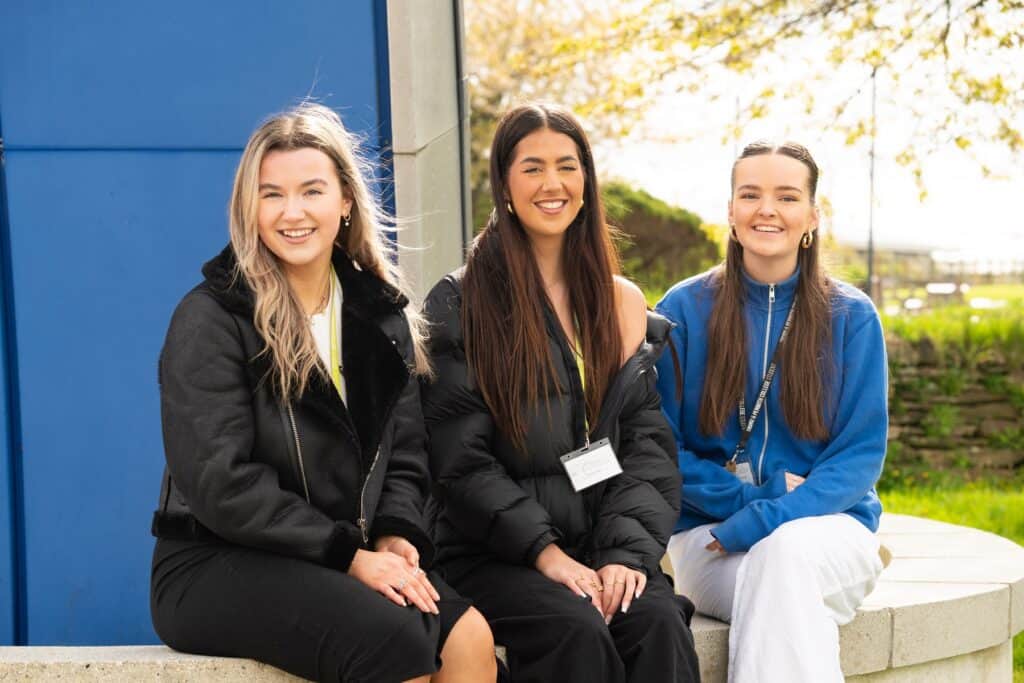 Three girls sit on a low wall outside on a bright day. They all smile and have coats on.