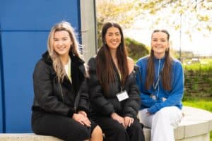 Three girls sit on a low wall outside on a bright day. They all smile and have coats on.