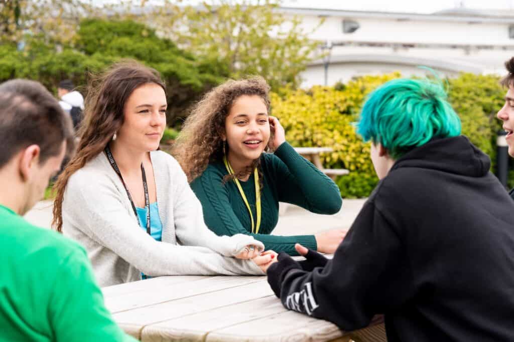 A group of students sit on a picnic bench outside. One student brushes her hair behind her ear and another crosses her arms on the table.