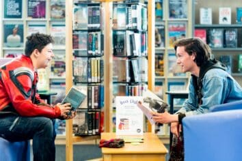 Two students look at each other with library bookshelves behind them. They both have books in their hands.