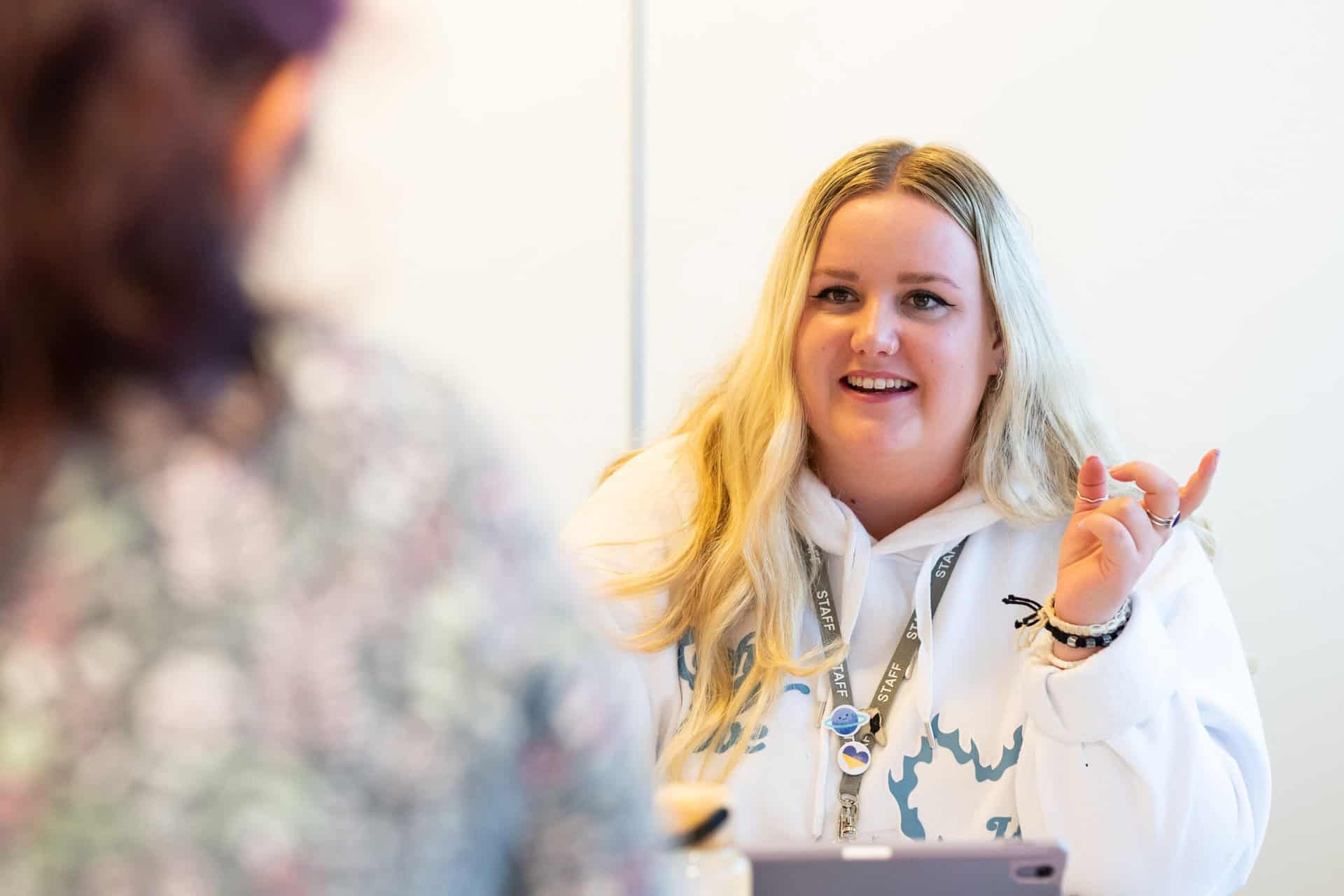 The camera focuses over the shoulder of an unknown person towards a member of staff who has blonde hair, a white jumper and is gesturing with her hand while smiling.