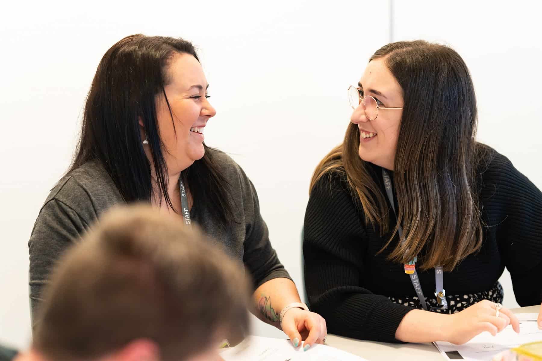 Two members of staff with dark hair and dark jumpers turn to each other and smile. They are both sat at a table.
