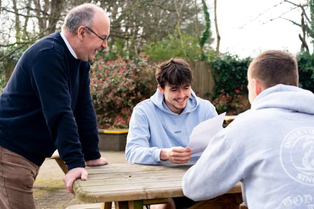 A tutor stands over two students who are sat on a picnic bench. One student holds a piece of paper and smiles.